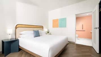 A modern hotel room with a bed, bedside tables, a nightstand, a wall-mounted lamp, and a door leading to a bathroom.