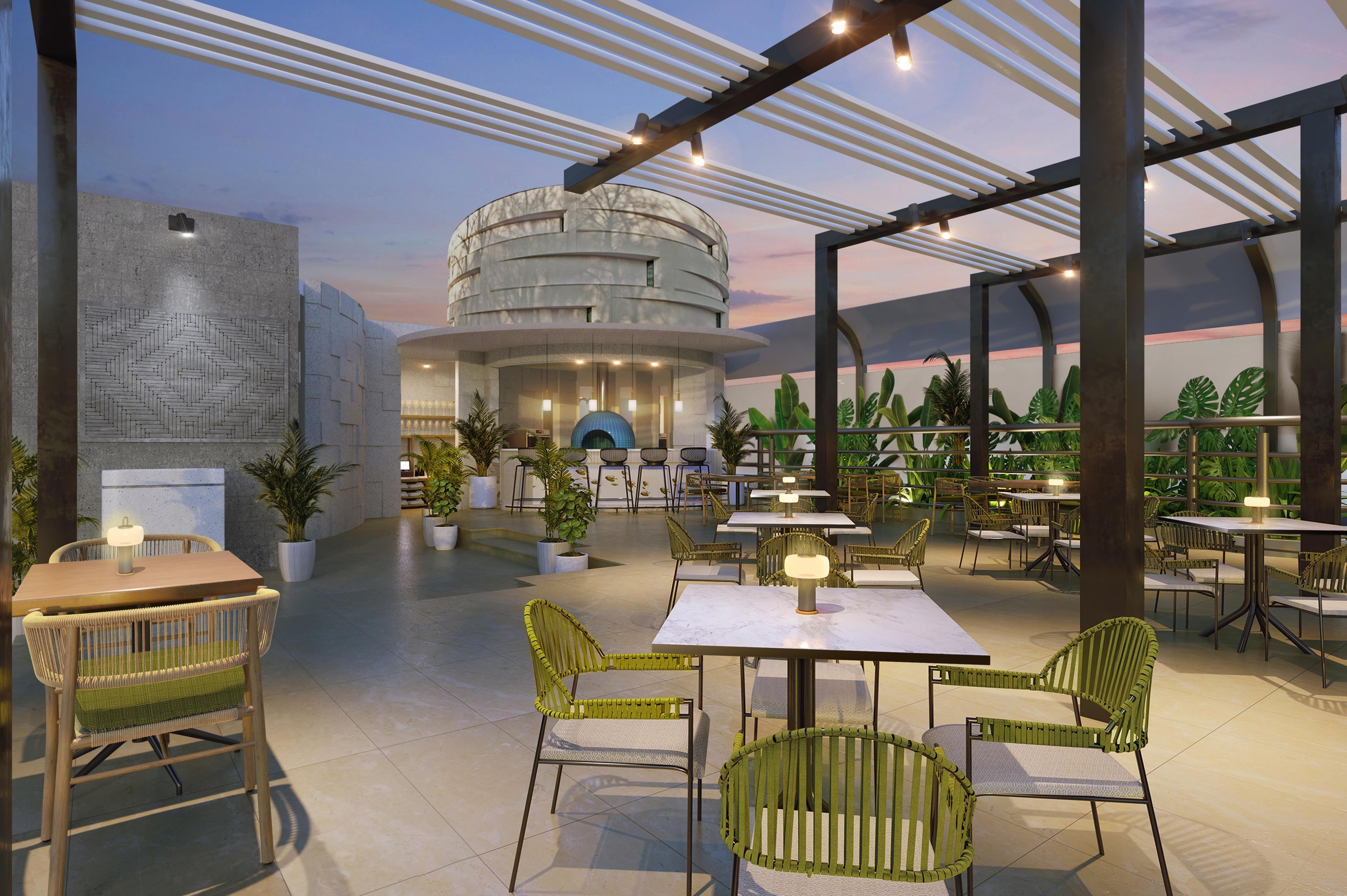 Un espacio de comedor al aire libre con mesas y sillas, un edificio circular al fondo y plantas en macetas.