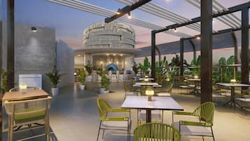 Un espacio de comedor al aire libre con mesas y sillas, un edificio circular al fondo y plantas en macetas.