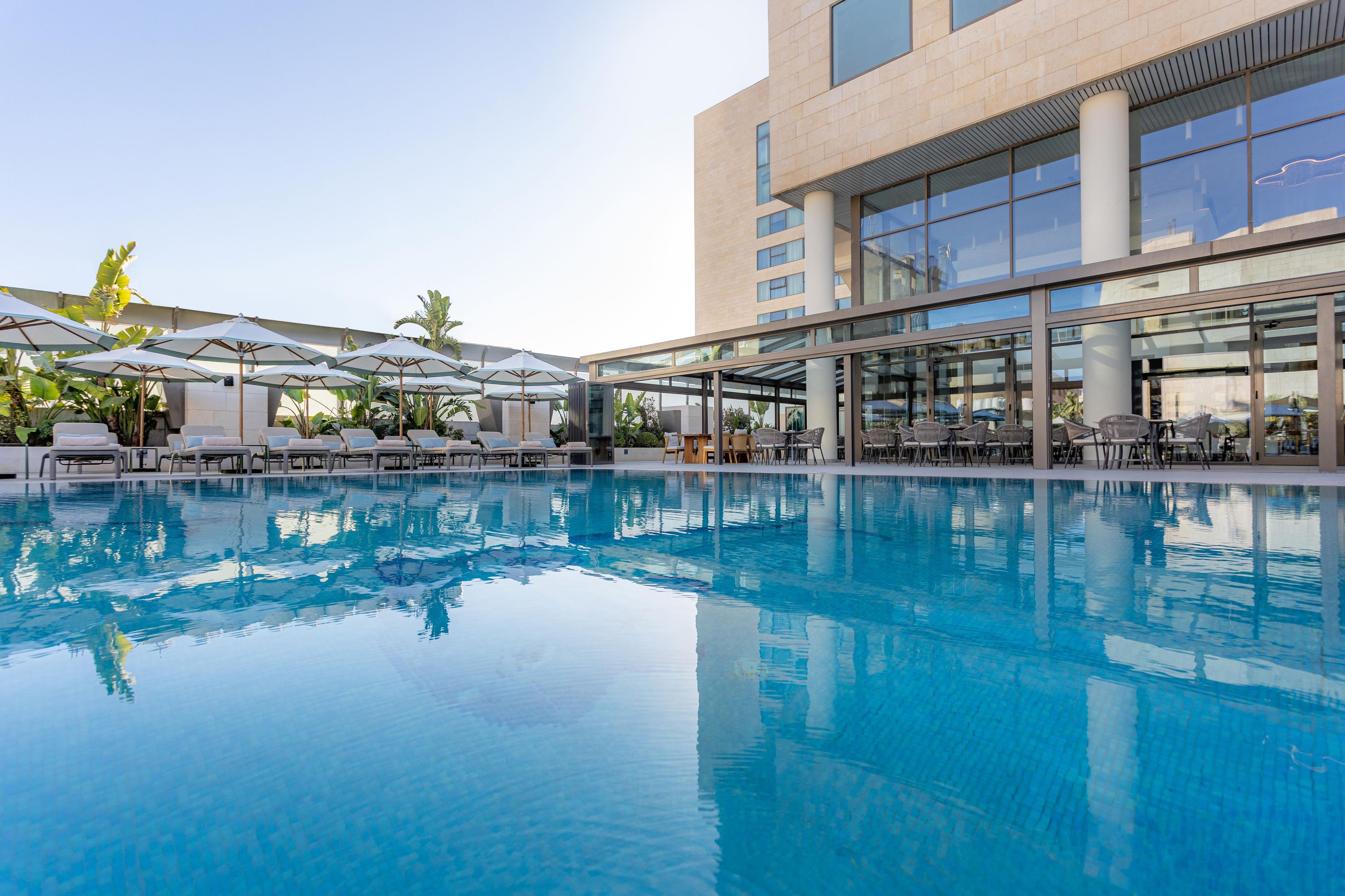 Una piscina con sillones de descanso y sombrillas, rodeada por un edificio moderno con grandes ventanales.