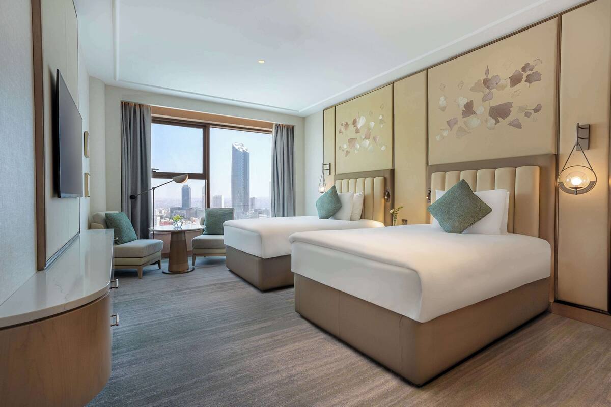 Ein Hotelzimmer mit zwei Betten, einem Schreibtisch, einem Stuhl, einem Fernseher und Blick auf die Stadt.