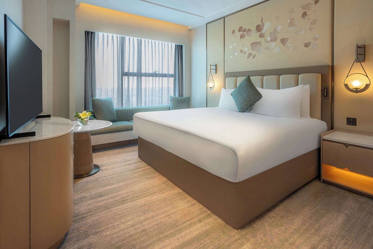 Ein Hotelzimmer mit einem großen Bett, einem Fernseher, einem kleinen Tisch mit einer Vase und einem Fenster mit Vorhängen.