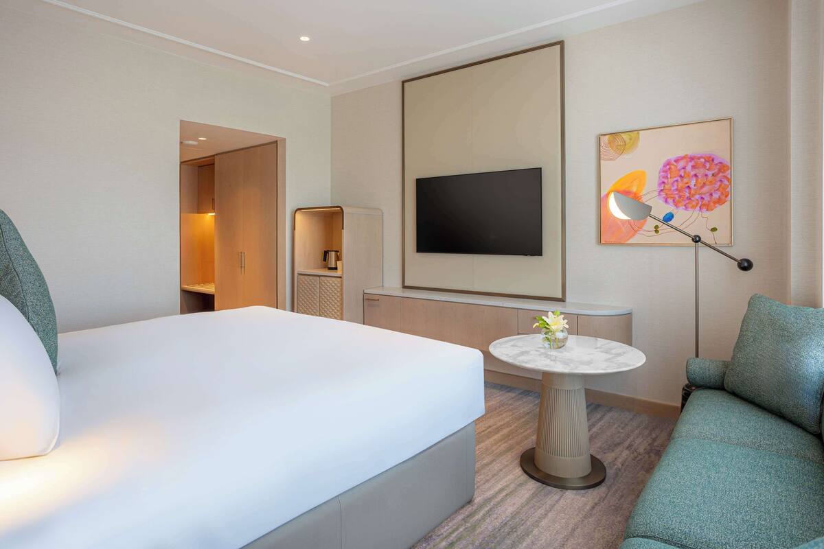 Ein Hotelzimmer mit einem großen Bett, einer Couch, einem kleinen runden Tisch mit einer Lampe, einem an der Wand befestigten Fernseher und einem Gemälde.