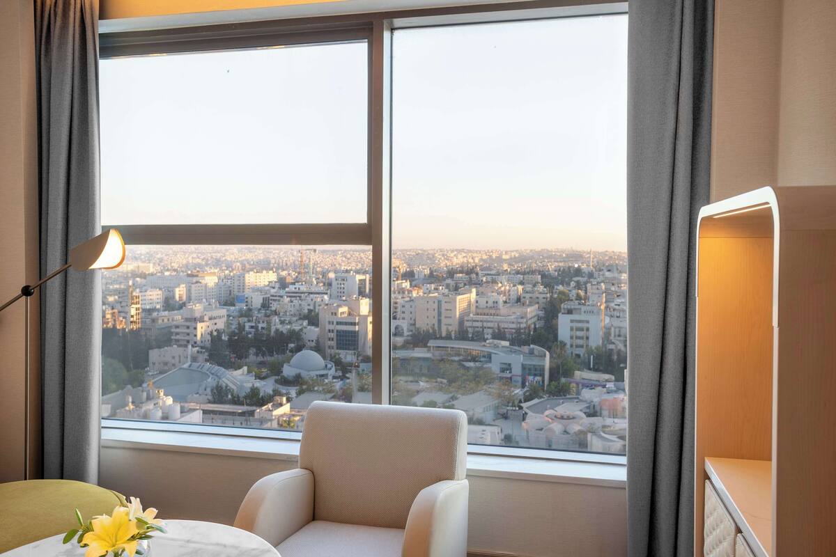Ein Zimmer mit großem Fenster und Blick auf die Stadt, ein Sessel, ein Tisch mit Blumenvase und eine Lampe.