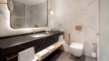A modern bathroom with a marble wall, a black countertop, a sink, a toilet, and a bench with towels.