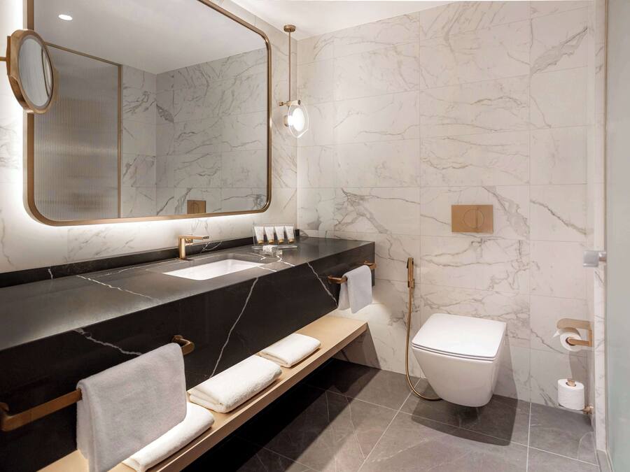 A modern bathroom with a marble wall, a black countertop, a sink, a toilet, and a bench with towels.