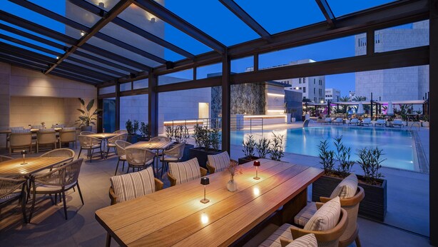 Un área de comedor al aire libre con vista a la piscina, con mesas y sillas de madera, plantas en macetas y un diseño arquitectónico moderno.