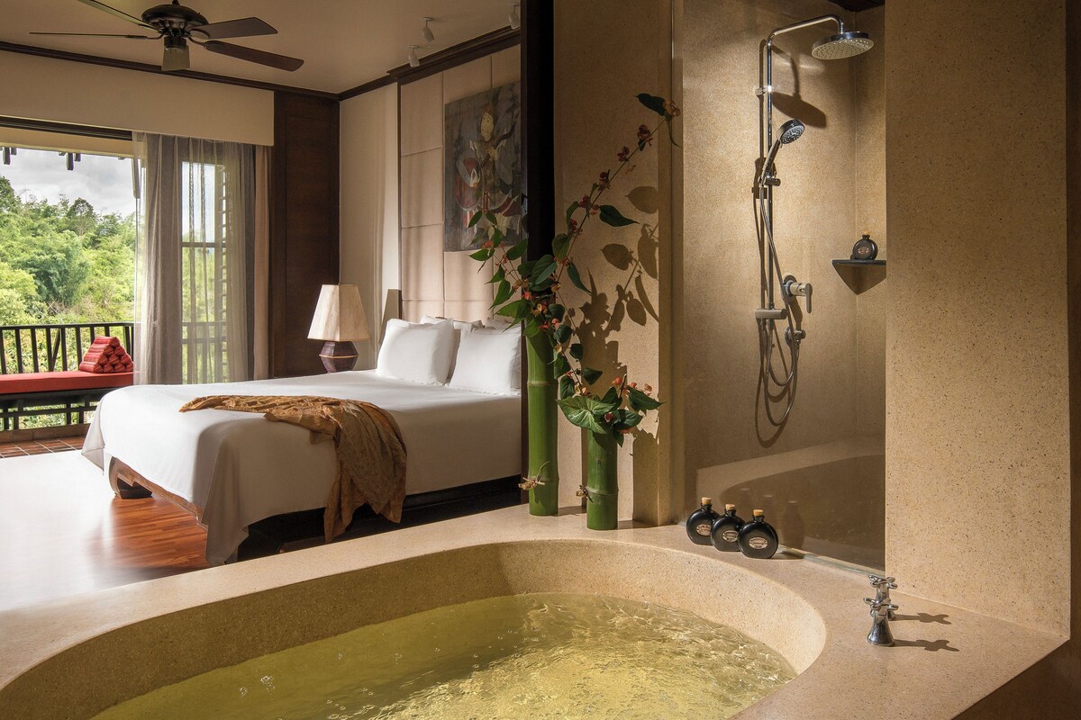 A bedroom with a large bed, a bathroom with a bathtub, and a balcony with a view of trees.