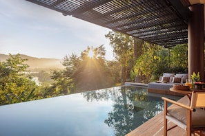 Una piscina infinita con vistas a un paisaje exuberante y colinas. Hay una zona de estar con un sofá y una mesa con una cesta y una planta.