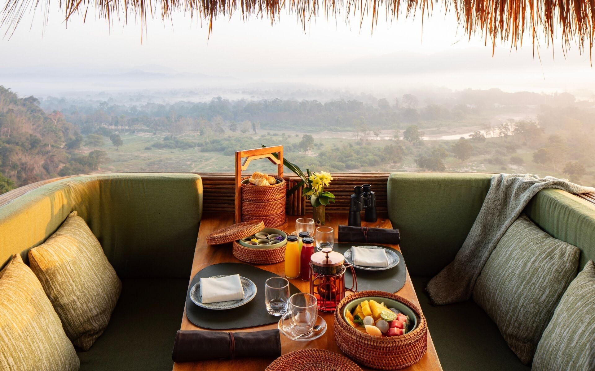 A dining setup with a table, plates, glasses, a fruit bowl, and a basket of bread, overlooking a scenic landscape.