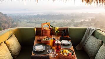 A dining setup with a table, plates, glasses, a fruit bowl, and a basket of bread, overlooking a scenic landscape.