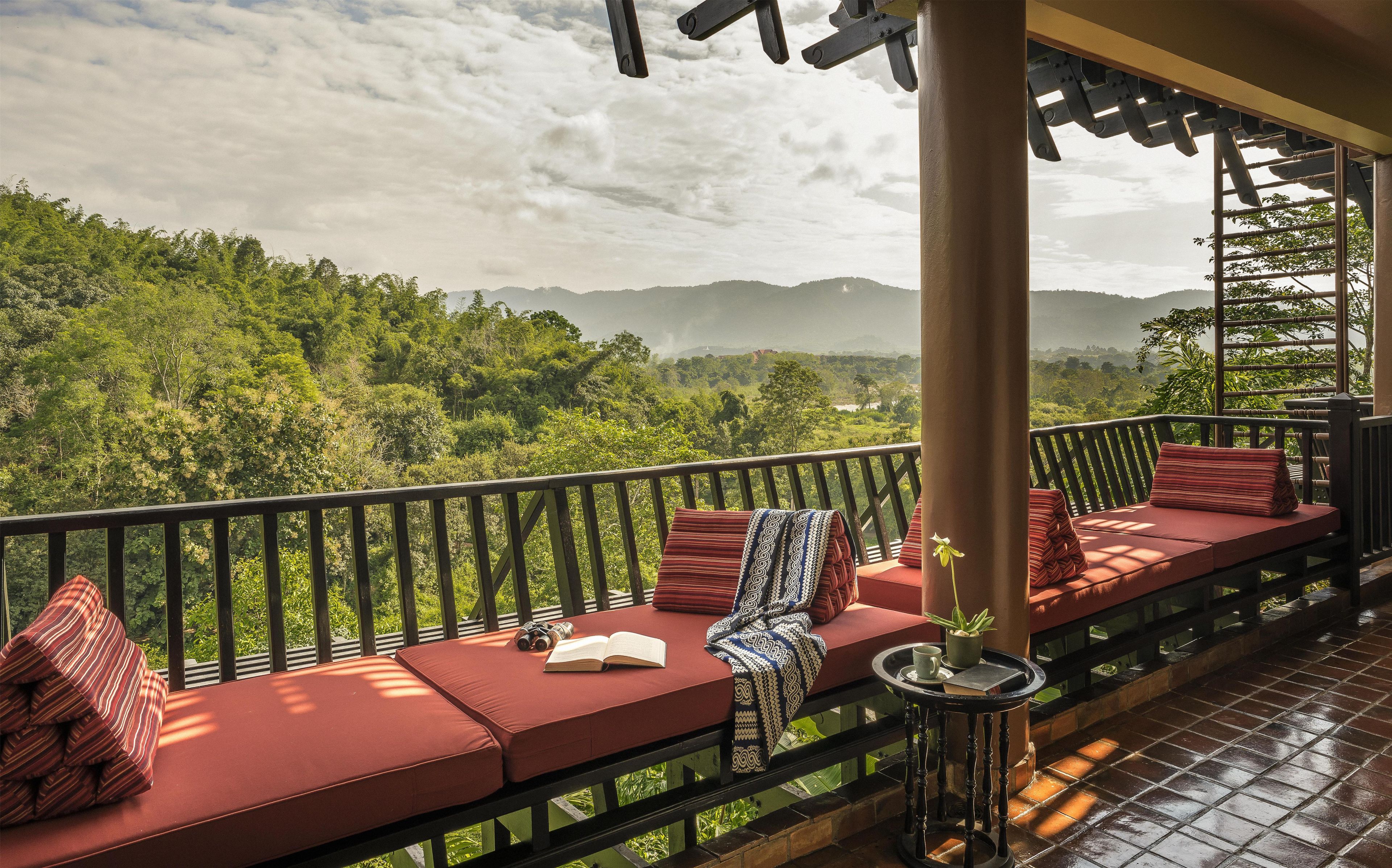 A balcony with red cushioned seating, a small table with a book and a cup, and a view of a lush green landscape with mountains in the distance.