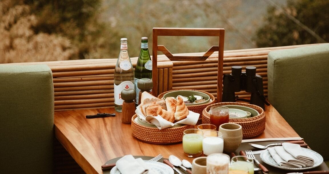 Mesa de comedor dispuesta con comida, bebidas y salsas, rodeada de asientos acolchados.