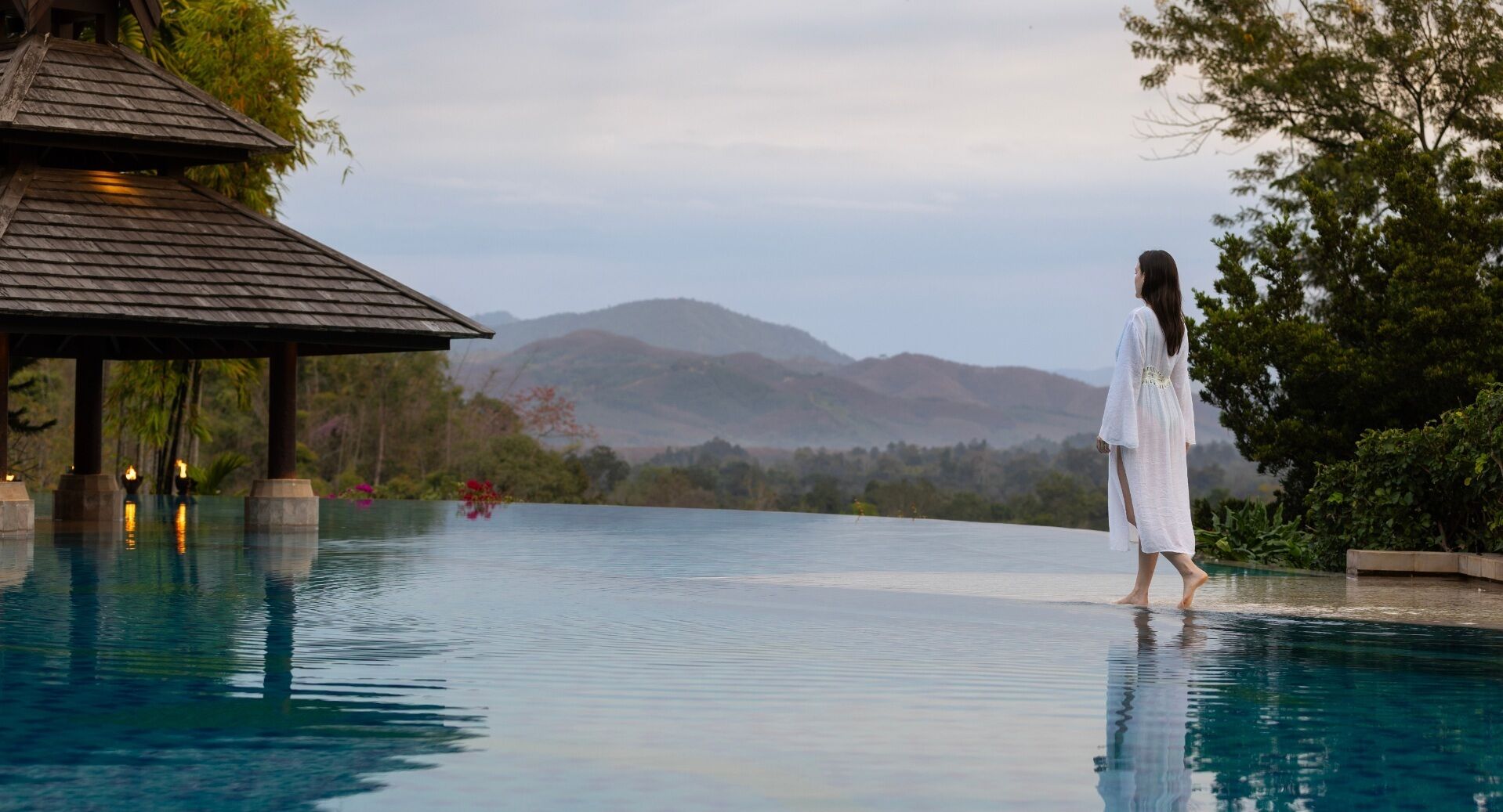 A person standing at the edge of a pool with a view of mountains and a gazebo in the background.