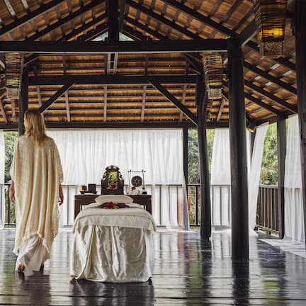 A covered outdoor area with a bed and a table, wooden beams, and hanging lanterns.