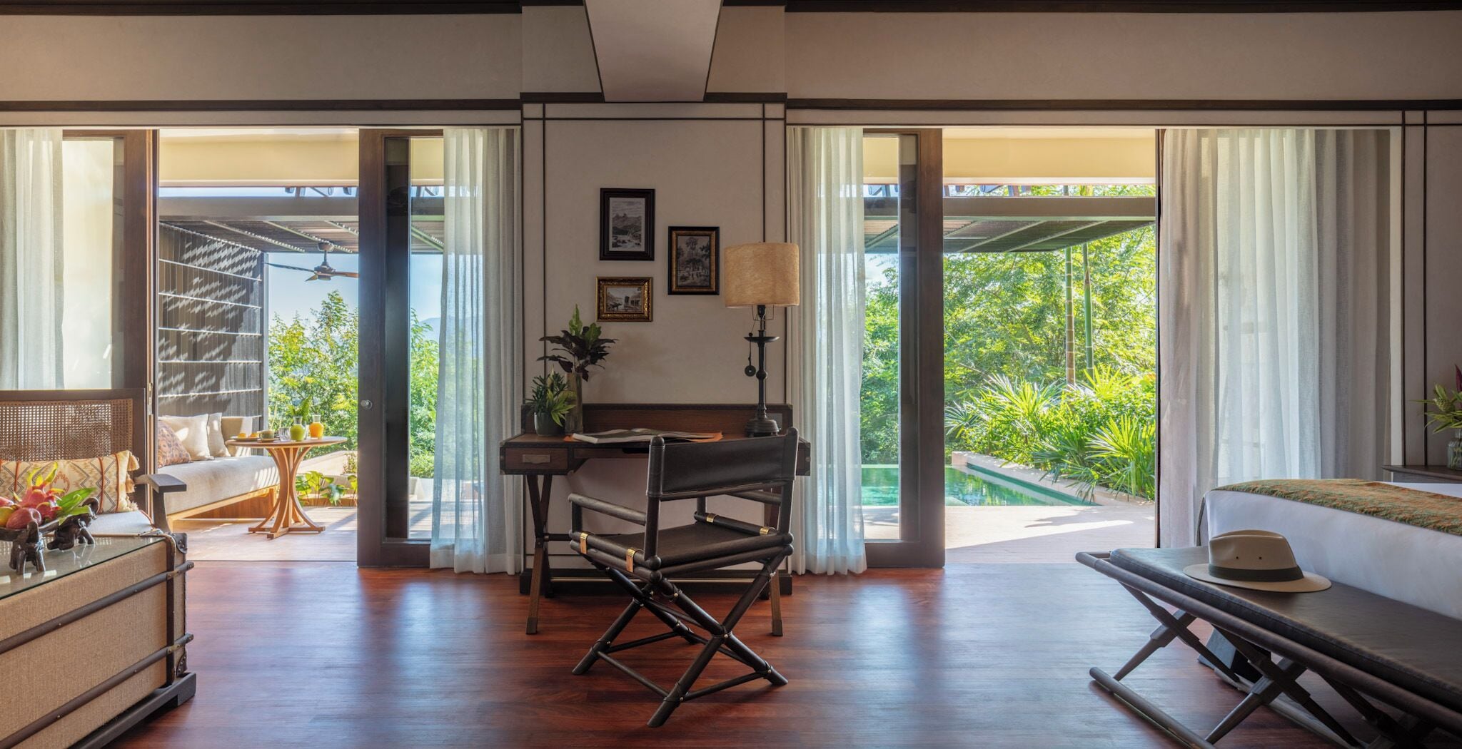 A spacious room with a wooden floor, a desk with a chair, a sofa, and a bed. There is a window with curtains, a view of greenery outside, and a small table with a lamp.