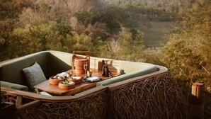 A dining area with a table set for two, surrounded by wicker furniture, overlooking a scenic landscape with hills and trees. - Anantara Golden Triangle Elephant Camp & Resort (Chiang Saen)