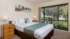A bedroom with a large window, a bed with a patterned bedspread, a nightstand with a lamp, and a painting on the wall.