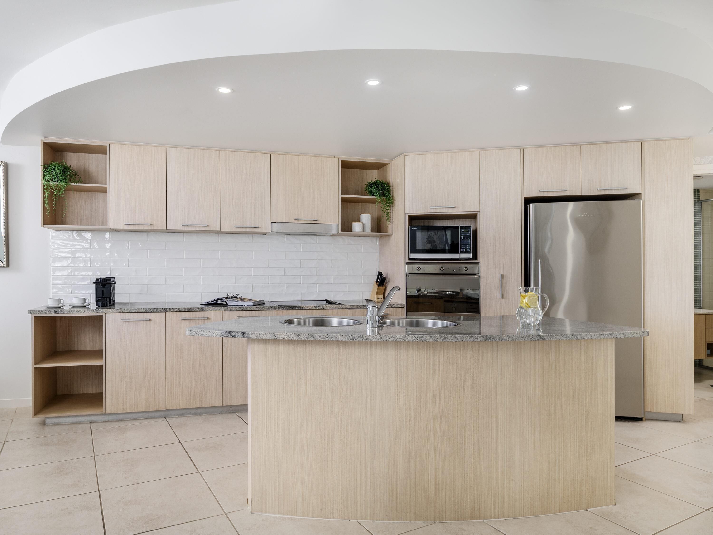 A modern kitchen with light wood cabinets, a central island, stainless steel appliances, and a white tiled backsplash.