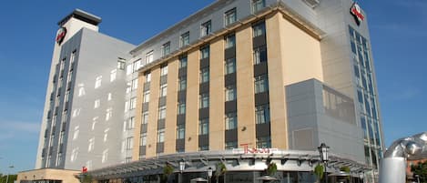 Un hotel moderno con fachada plana, múltiples ventanas y una zona de estar exterior cubierta con mesas y sillas.