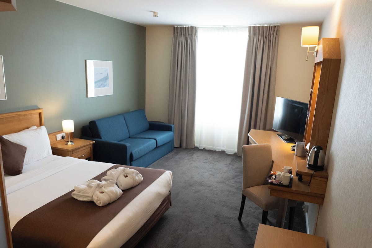 A hotel room with a bed, a blue sofa, a desk with a chair, a TV, and a window with curtains.