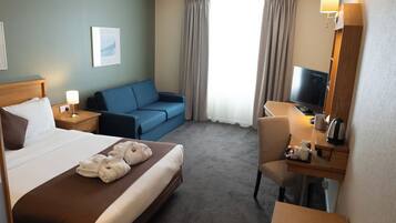 Habitación de hotel con cama, sofá azul, escritorio con silla, televisión y ventana con cortinas.