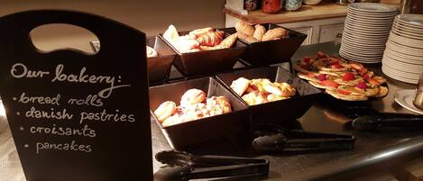 A bakery counter with various baked goods like bread rolls, Danish pastries, croissants, and pancakes.
