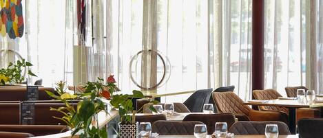A restaurant with booths and tables, large windows with sheer curtains, and hanging pendant lights.