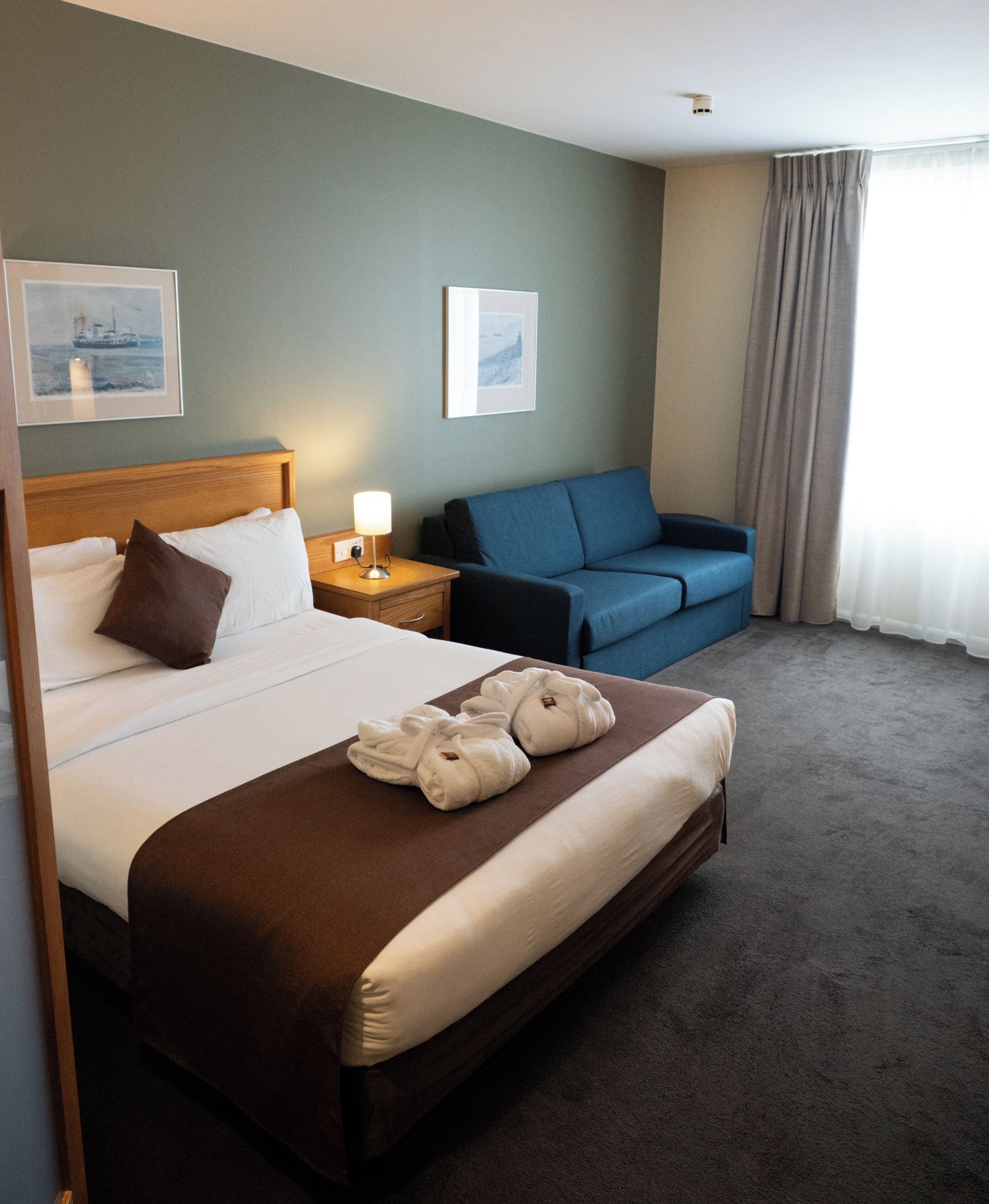 A hotel room with a large bed, a bedside table with a lamp, a blue sofa, and a window with curtains.