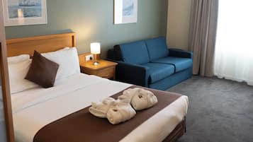 A hotel room with a large bed, a bedside table with a lamp, a blue sofa, and a window with curtains.