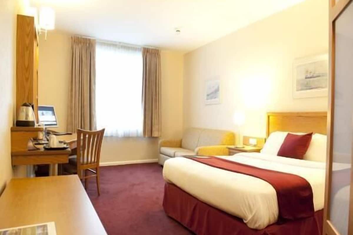 A hotel room with a bed, desk, chair, and a window with curtains.