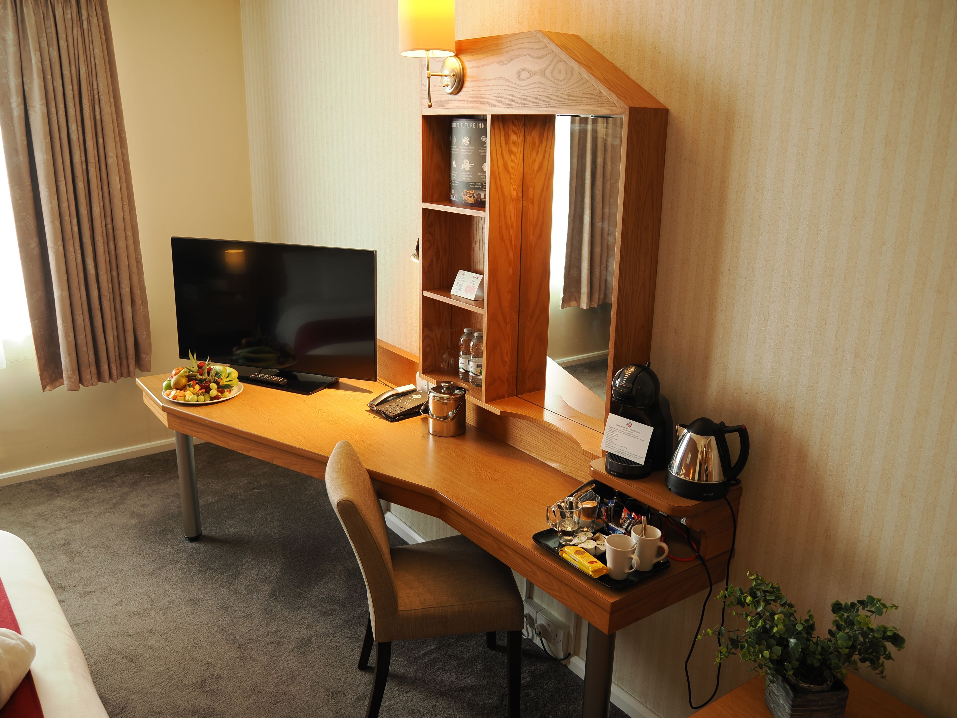 A hotel room with a wooden desk, a television, a minibar, a plant, and a window with curtains.
