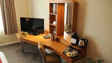 A hotel room with a wooden desk, a television, a minibar, a plant, and a window with curtains.