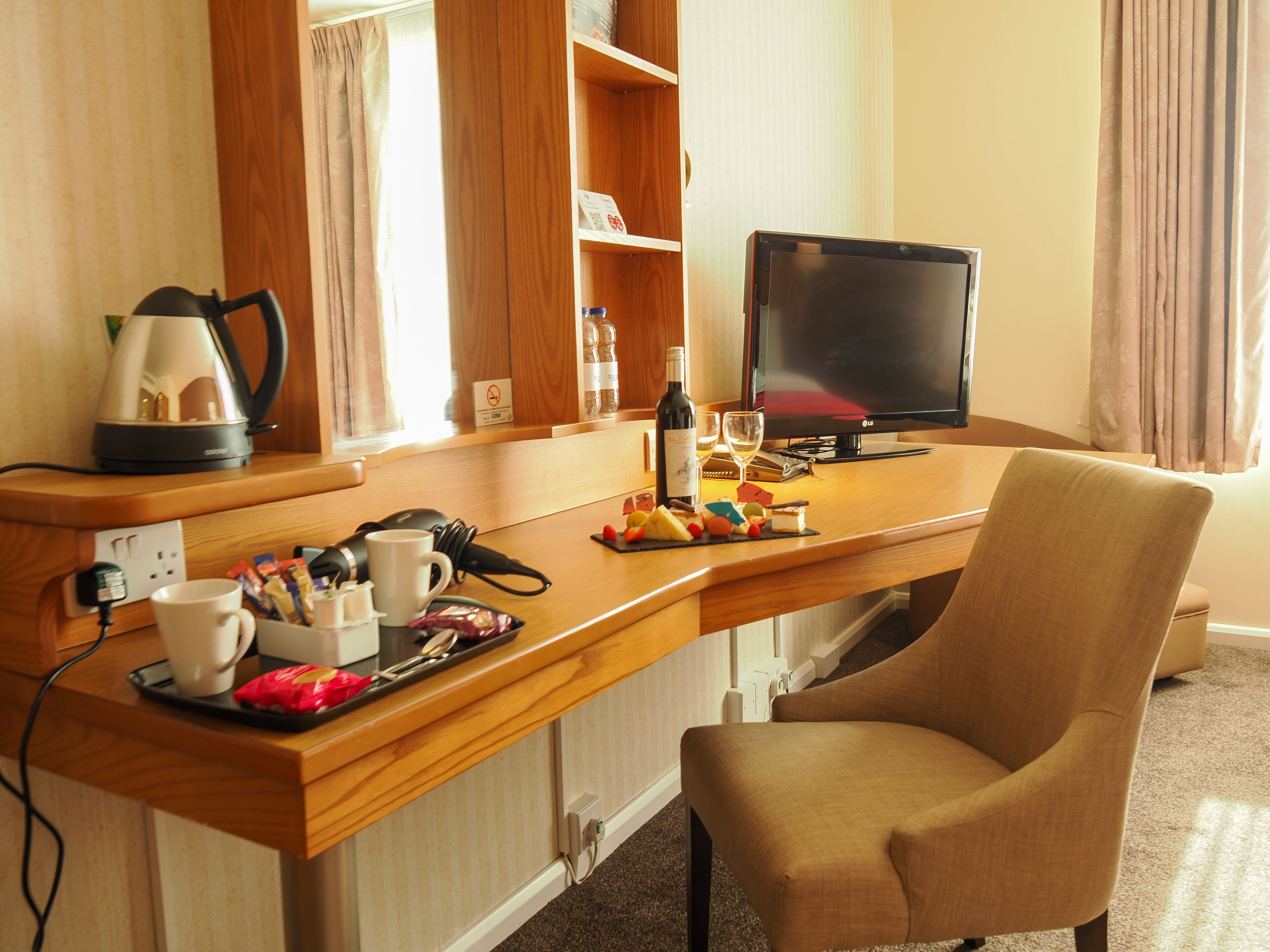 A hotel room with a wooden desk, a television, a chair, and various items like a kettle, wine bottle, and snacks.