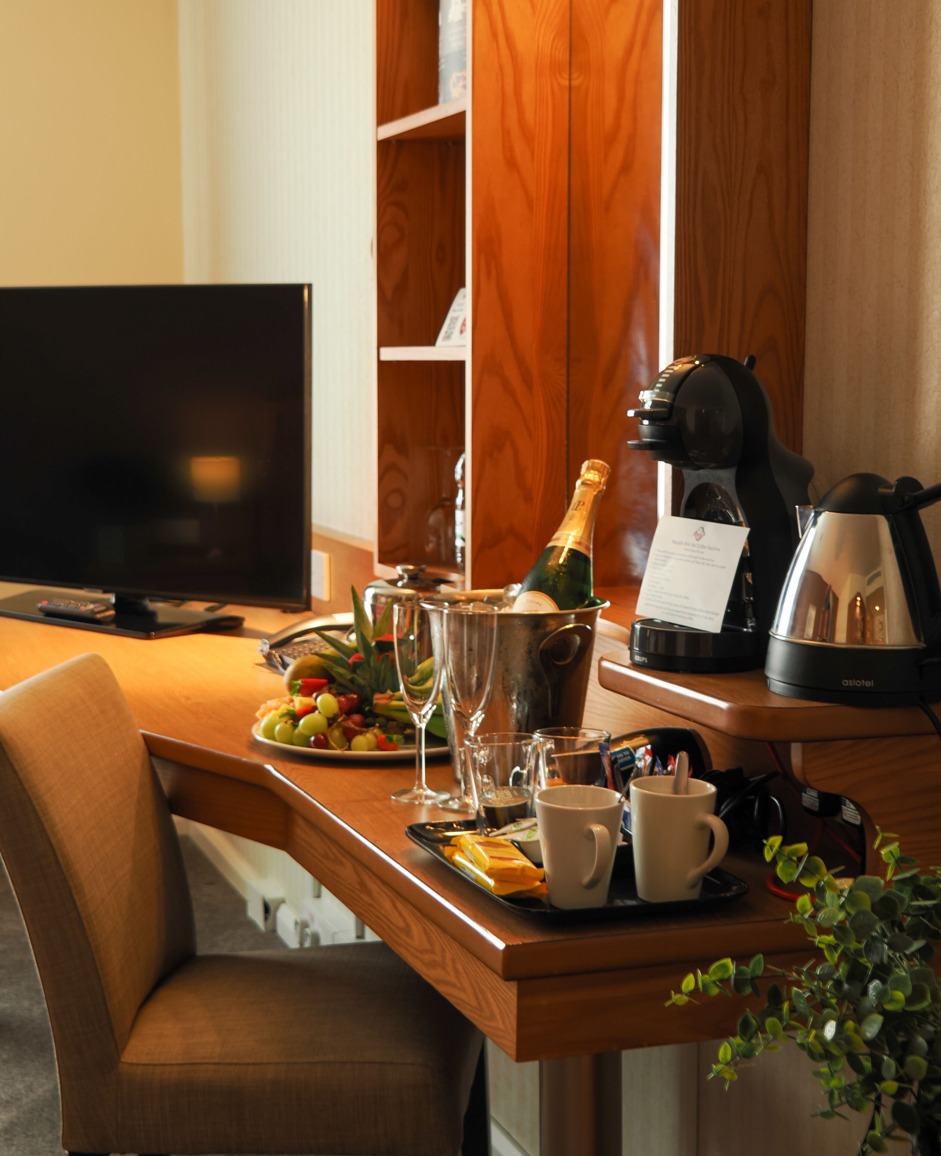 Habitación de hotel con escritorio de madera, televisor, una botella de champán, cafetera y una bandeja de fruta.
