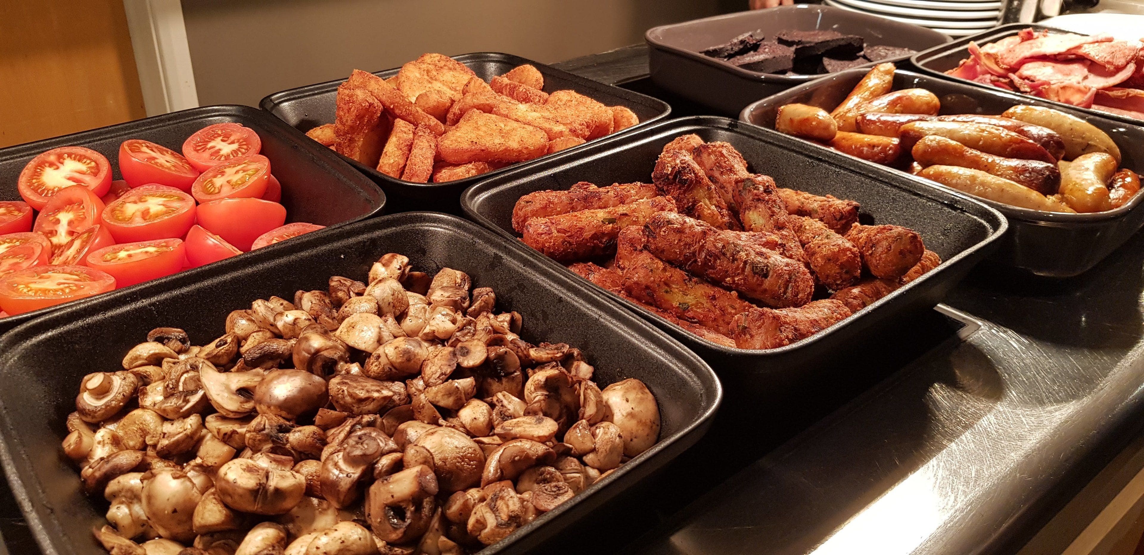 A buffet with various food items including sliced tomatoes, mushrooms, and sausages.