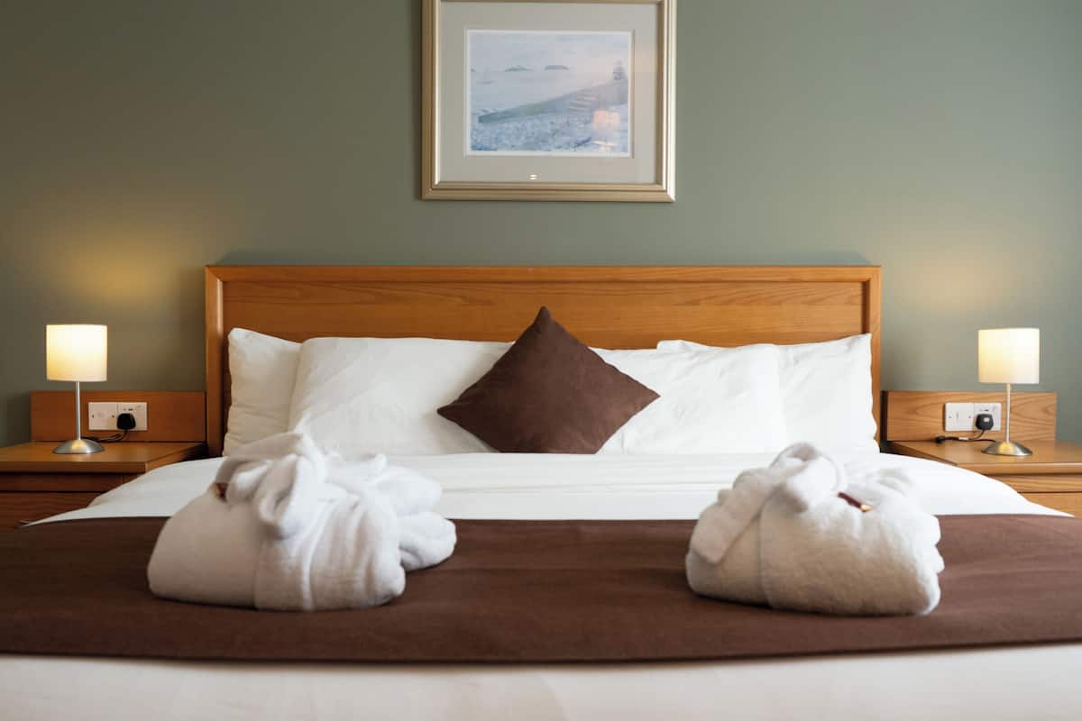 A hotel room with a wooden headboard, a framed picture on the wall, two bedside lamps, and two towels on the bed.