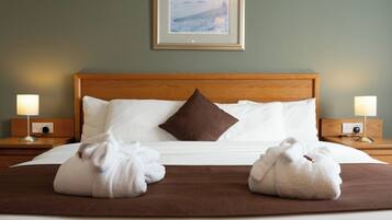 Quarto de hotel com cabeceira de madeira, quadro na parede, duas luminárias de apoio e duas toalhas sobre a cama.