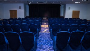A conference room with rows of chairs, a carpeted floor, a stage, and a projector screen.
