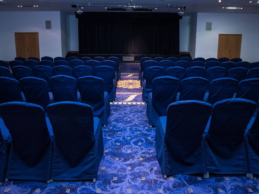 Un salón de conferencias con filas de sillas, suelo alfombrado, un escenario y una pantalla de proyector.