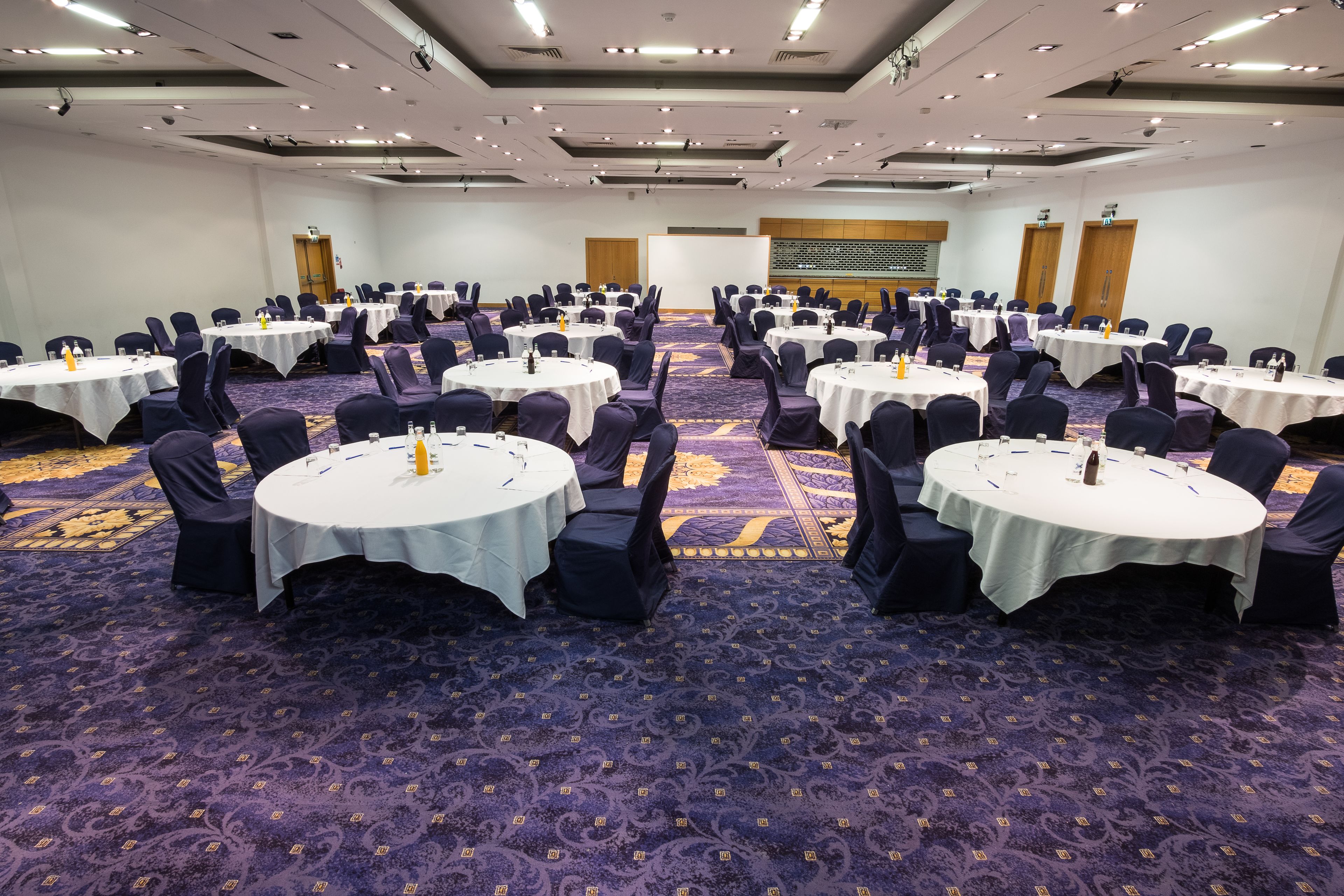 A conference room with multiple round tables, each with a white tablecloth and black chairs. The room has a carpeted floor and a high ceiling with recessed lighting.