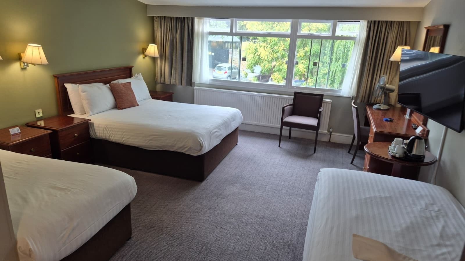 A hotel room with two beds, a desk, a chair, a television, and a window with a view of trees.