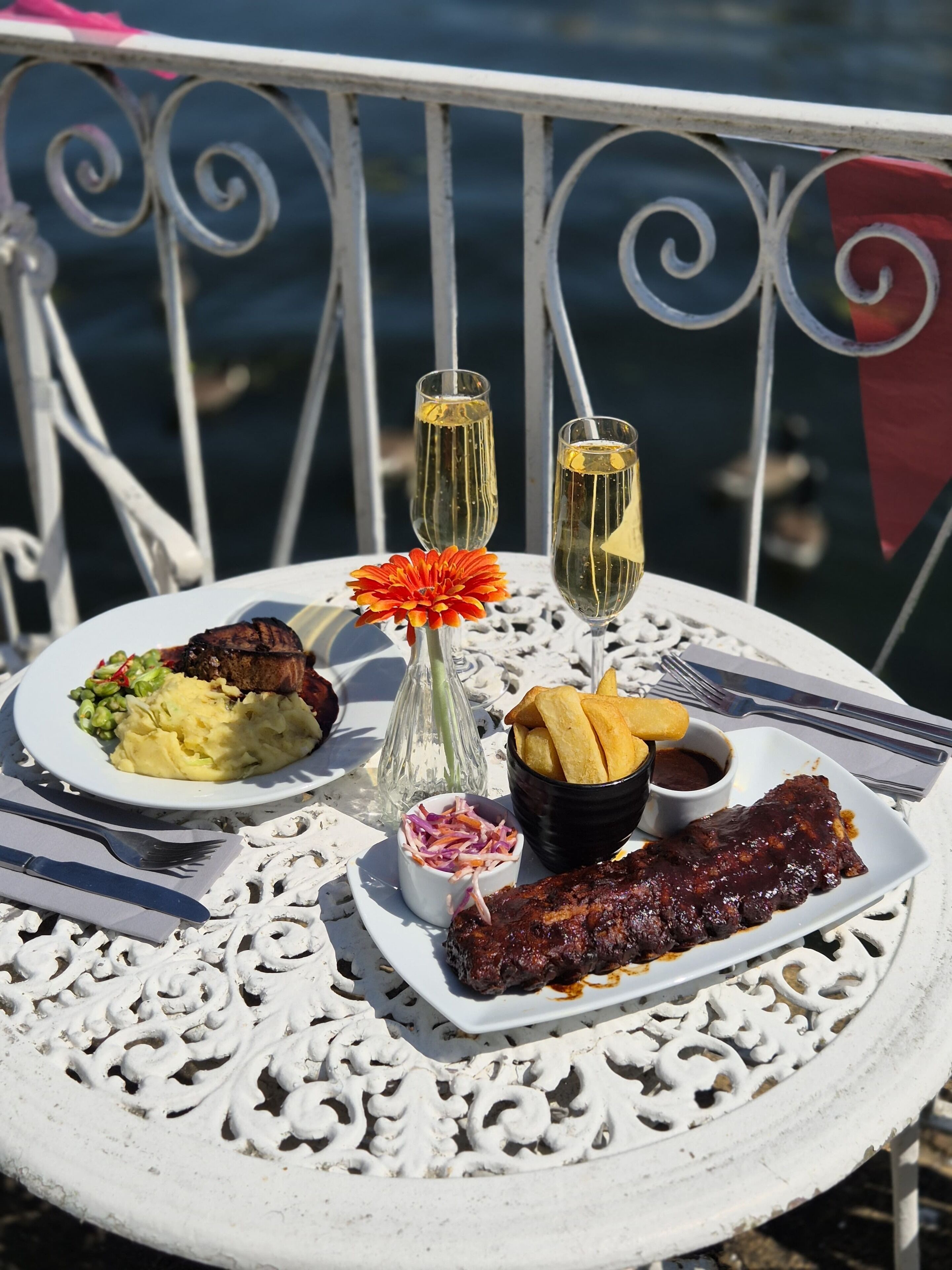 Una mesa con comida que incluye carne, puré de papas, ensalada de repollo y papas fritas, acompañada de dos vasos de una bebida amarilla, probablemente champán, sobre una mesa blanca decorada al aire libre.