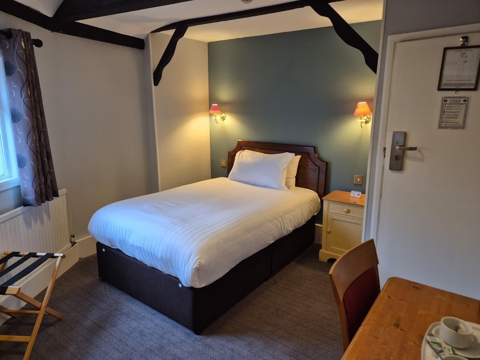 A bedroom with a bed, bedside lamps, a wooden nightstand, a chair, and a table with a cup and saucer.