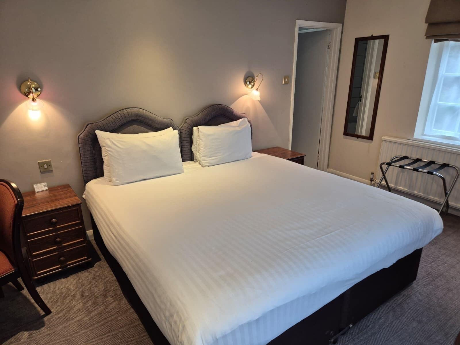 A neatly made bed with white linens, a wooden nightstand with a drawer, a chair, and two wall-mounted lamps.