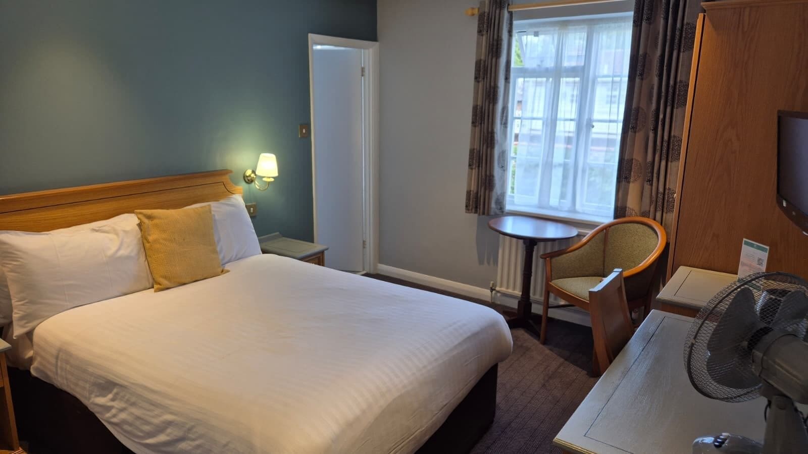 A hotel room with a large bed, a small table, a chair, a desk, a lamp, and a television.