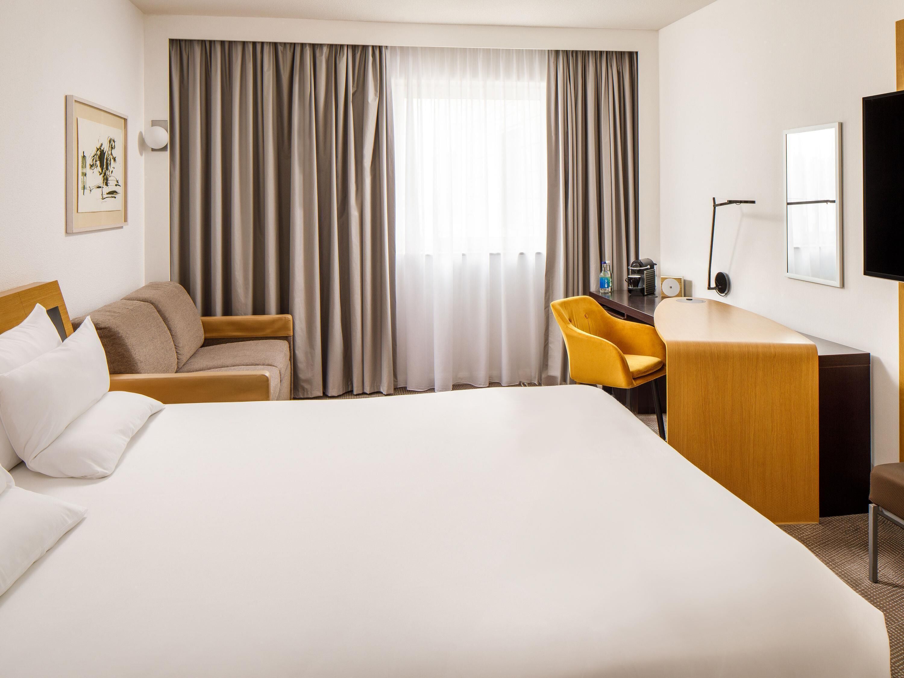 Una camera d'albergo con un letto, una scrivania, una sedia, un televisore e una finestra con tende.