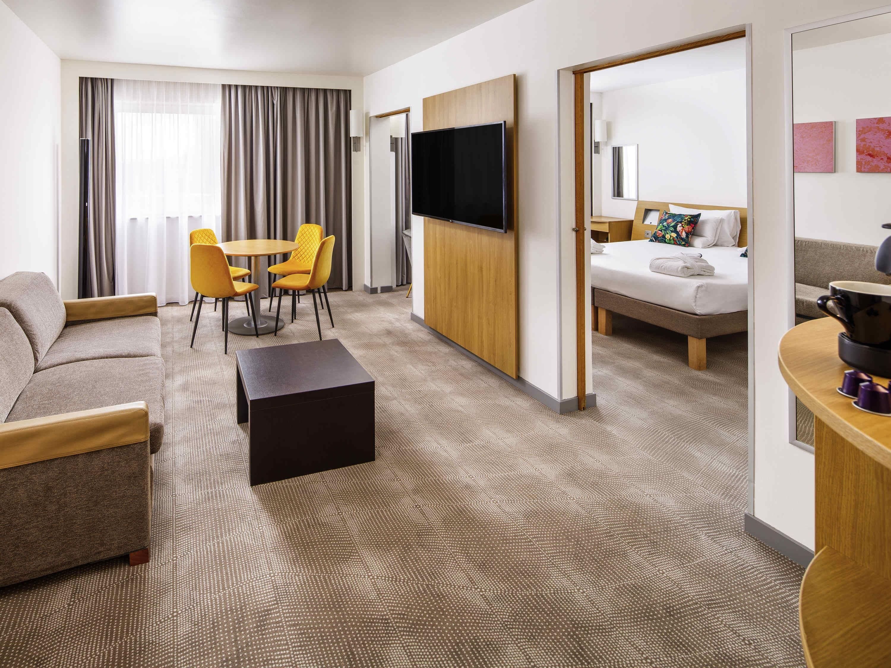 Ein Hotelzimmer mit Sofa, einem Esstisch mit Stühlen, einem an der Wand montierten Fernseher, einem Bett und einer kleinen Küchenzeile mit Kaffeemaschine.