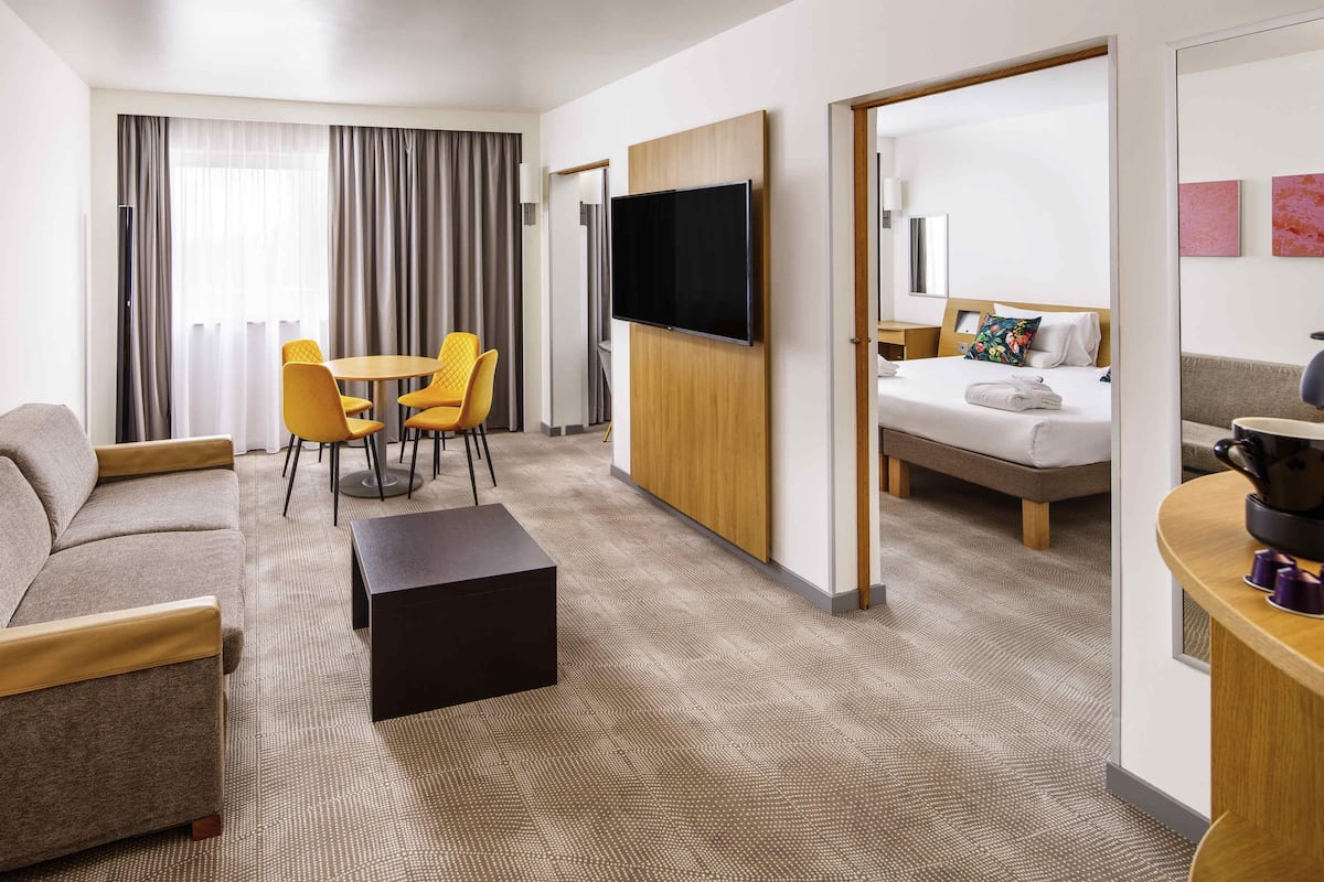 A hotel room with a sofa, a dining table with chairs, a TV mounted on the wall, a bed, and a small kitchen area with a coffee maker.