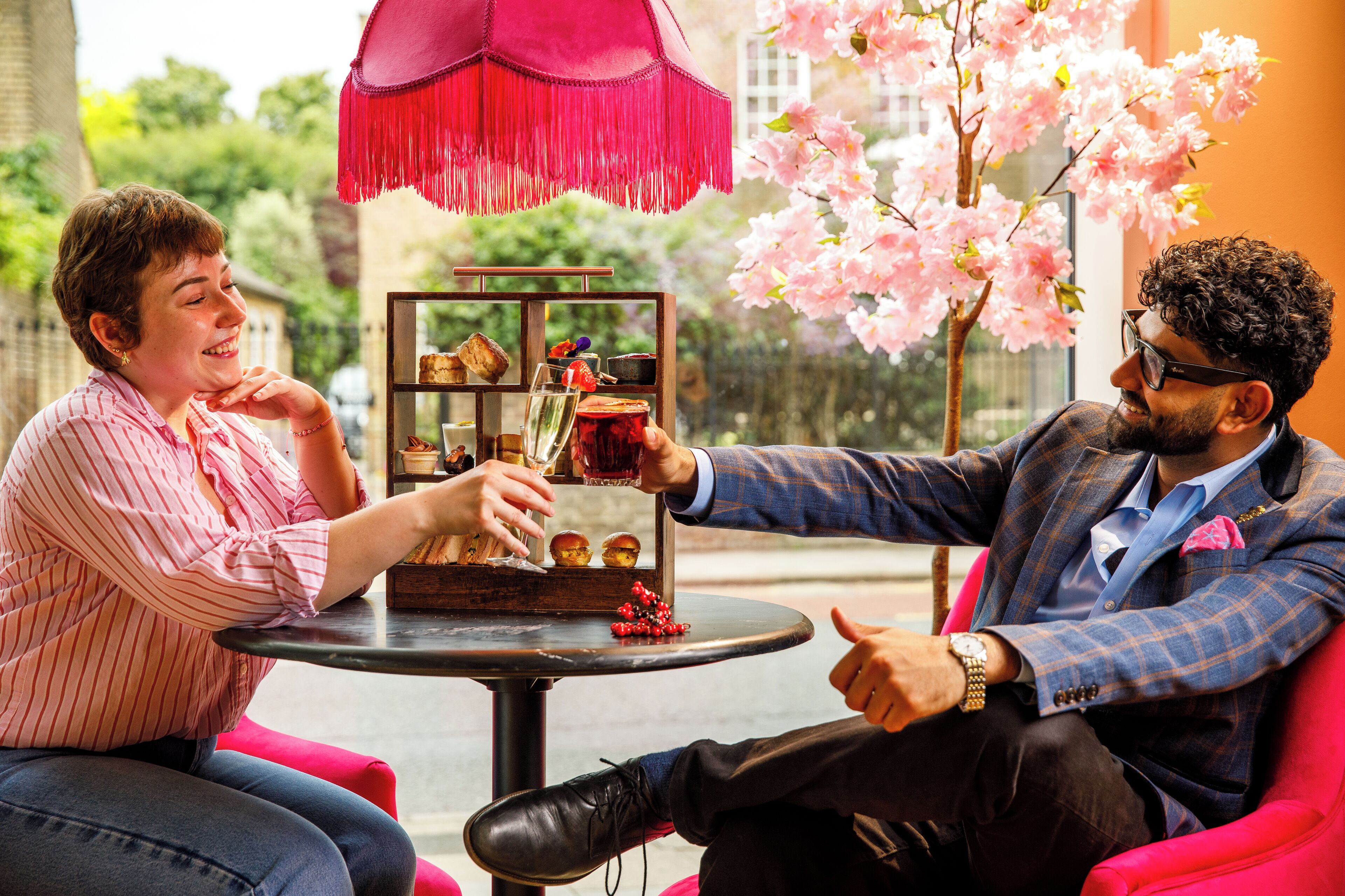 Zwei Personen sitzen an einem Tisch mit einer Dessertauslage, einem pinken Sonnenschirm und Kirschblüten im Hintergrund.